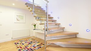 A modern staircase with glass railings and wooden steps, a colorful rug, and a small table with a vase of flowers. - Shamal (St Ives)