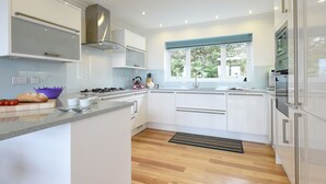 A modern kitchen with white cabinetry, a gas stove, a sink, and a window with a view of greenery. - Shamal (St Ives)