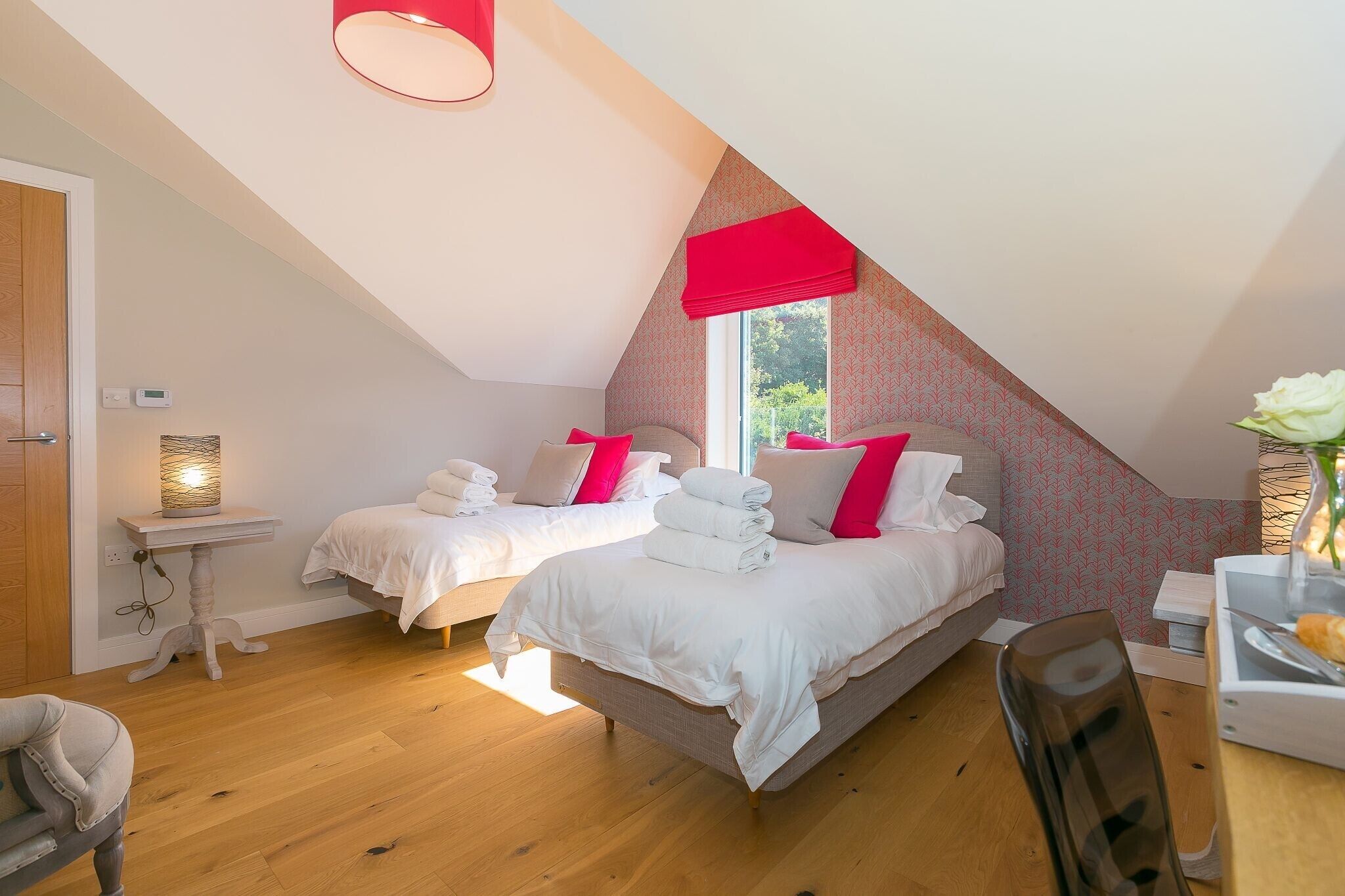 A bedroom with two beds, a small table, a chair, and a window with a red valance.