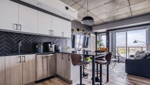 Et moderne køkken med hvide skabe, en backsplash i sort fiskebensmønster og stålstål-hvidevarer. Der er et spiseområde med et bord og barstole, samt et stort vindue med udsigt over byen.