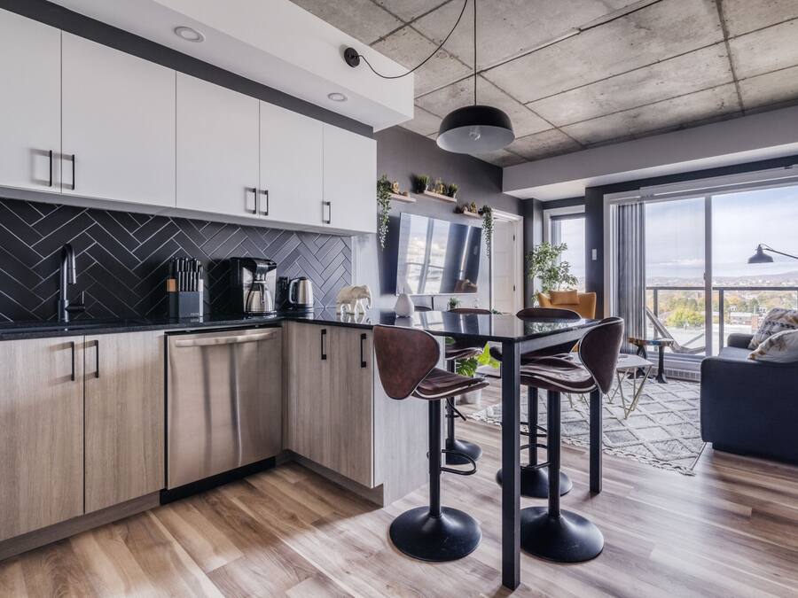 Una cocina moderna con armarios blancos, salpicadero tipo espiga en color negro y electrodomésticos de acero inoxidable. Cuenta con un comedor con mesa y taburetes de bar, y un amplio ventanal con vistas a la ciudad.