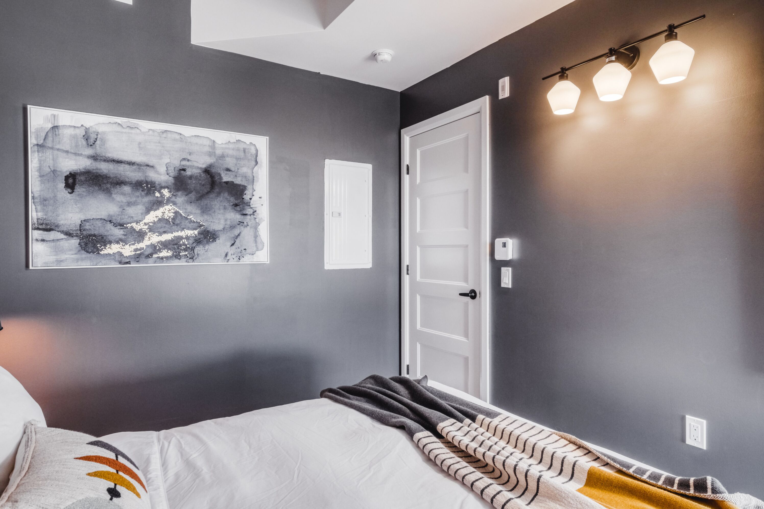 A bedroom with a bed, a large abstract painting, a white door, and a wall-mounted light fixture.