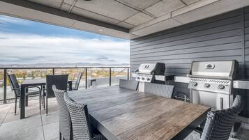 Um pátio externo com mesa e cadeiras de madeira, duas churrasqueiras a gás e vista para a paisagem urbana.