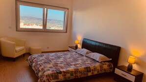 Un dormitorio con cama, dos lámparas de noche, una silla y una ventana con vistas a edificios y colinas.