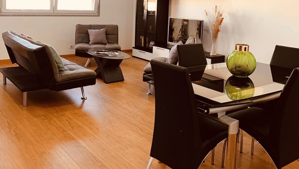 Una sala moderna con sofá, mesa de centro y sillones. También hay una mesa de comedor con sillas. El piso es de madera y cuenta con un ventanal grande.