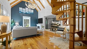 Ein geräumiges Wohnzimmer mit hoher Decke, Holzbalken, einer Treppe und einem durch eine offene Tür einsehbaren Küchenbereich.