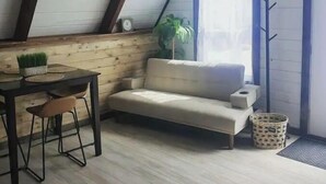 Et hyggeligt loftrum med træloft, en hvid sofa, et lille spisebord med stole, en potteplante og et knagered.