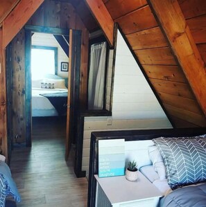 Une chambre confortable située sous les toits, avec un plafond en pente, des poutres en bois, un lit agrémenté d’un coussin à motifs gris et blancs, et une petite plante posée sur la table de chevet.