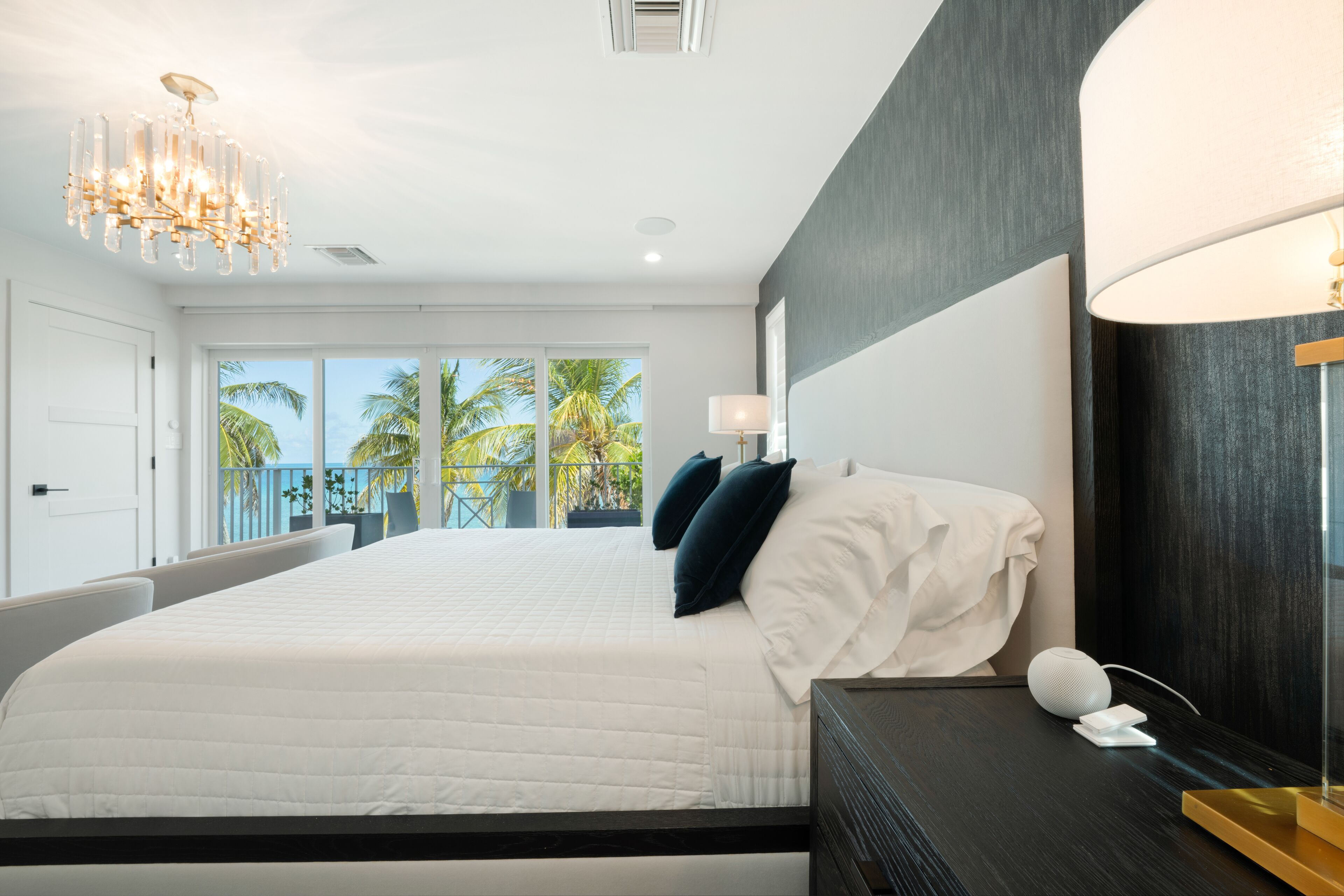 Een moderne slaapkamer met een groot bed, een kroonluchter, een bedlampje en uitzicht op palmbomen door grote ramen.