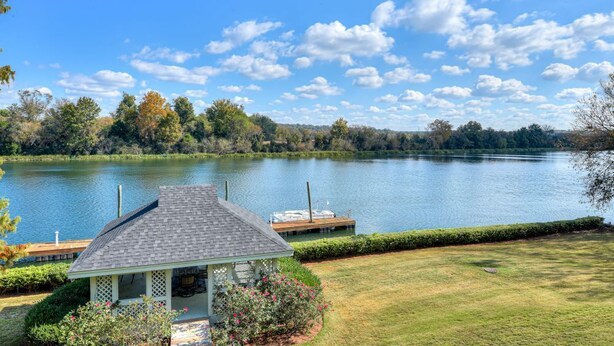 A lakeside gazebo with a dock extending into a calm lake, surrounded by lush greenery and trees.