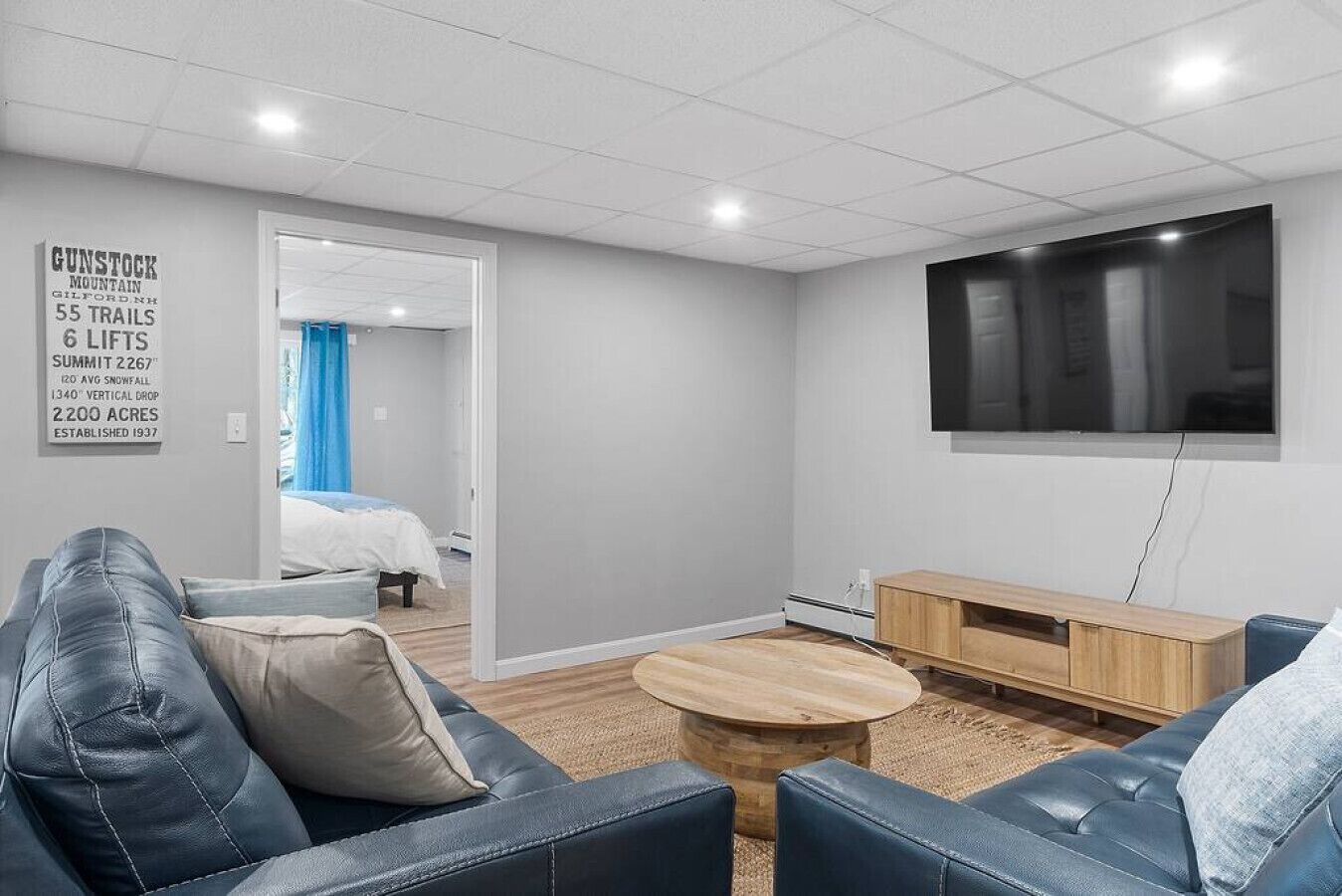 A modern living room with a large TV, leather sofas, a wooden coffee table, and a bed in the background.