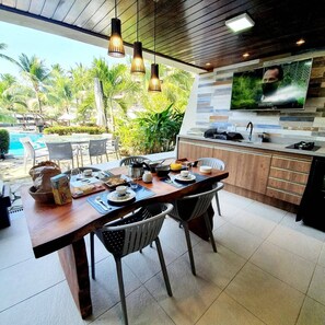 Cozinha moderna com área de jantar, TV montada acima do balcão e vista para uma piscina e área verde lá fora.