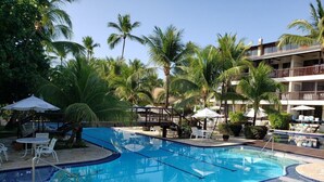 A swimming pool with lounge chairs and umbrellas, surrounded by palm trees and a building with balconies. - Flat Luxo Térreo Nannai Residence (Ipojuca)