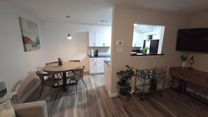 A modern kitchen with white cabinets, a dining area with a round table and chairs, and a small side table with a plant.