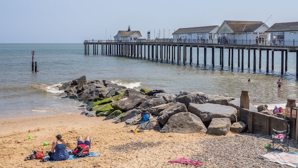 Een strand met een pier die zich in de zee uitstrekt, met een rij gebouwen en enkele mensen in het water.