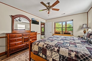 A bedroom with a wooden dresser, a large bed with a quilt, a ceiling fan, a window with a view, and a mirror.