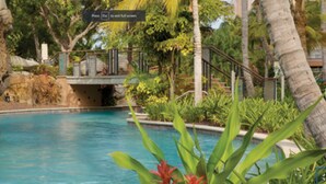 Un espace piscine agrémenté d’une formation rocheuse, d’un pont et de plantes tropicales.