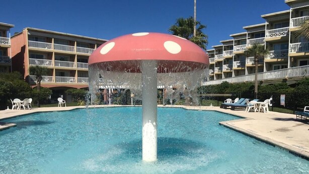 A swimming pool with a mushroom-shaped water feature in the center, surrounded by lounge chairs and a multi-story building with balconies. - Cute ocean view beach front condo w private balcony . Great LOCATION! Sleeps 6. (Galveston)