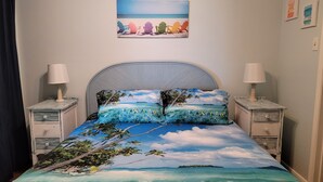 A bed with a tropical beach scene bedspread, two bedside lamps, a headboard, and a painting of beach chairs on the wall. - Cute ocean view beach front condo w private balcony . Great LOCATION! Sleeps 6. (Galveston)
