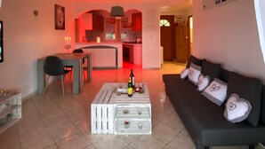 Ein Wohnzimmer mit einer Couch, einem Couchtisch und einem Essbereich mit Tisch und Stühlen. Im Hintergrund ist eine Küche erkennbar.