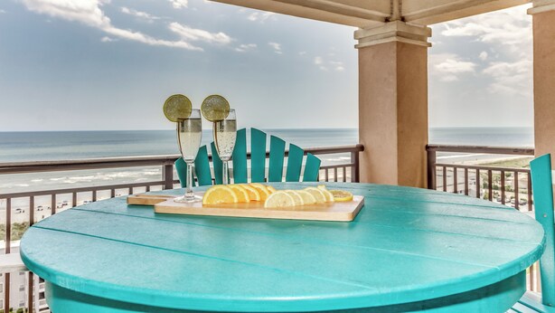 Ein türkisfarbener Tisch mit einem Glas Wasser, zwei Gläsern mit Limettenscheiben und einem Holzbrett mit Zitronenscheiben auf einem Balkon mit Blick aufs Meer.
