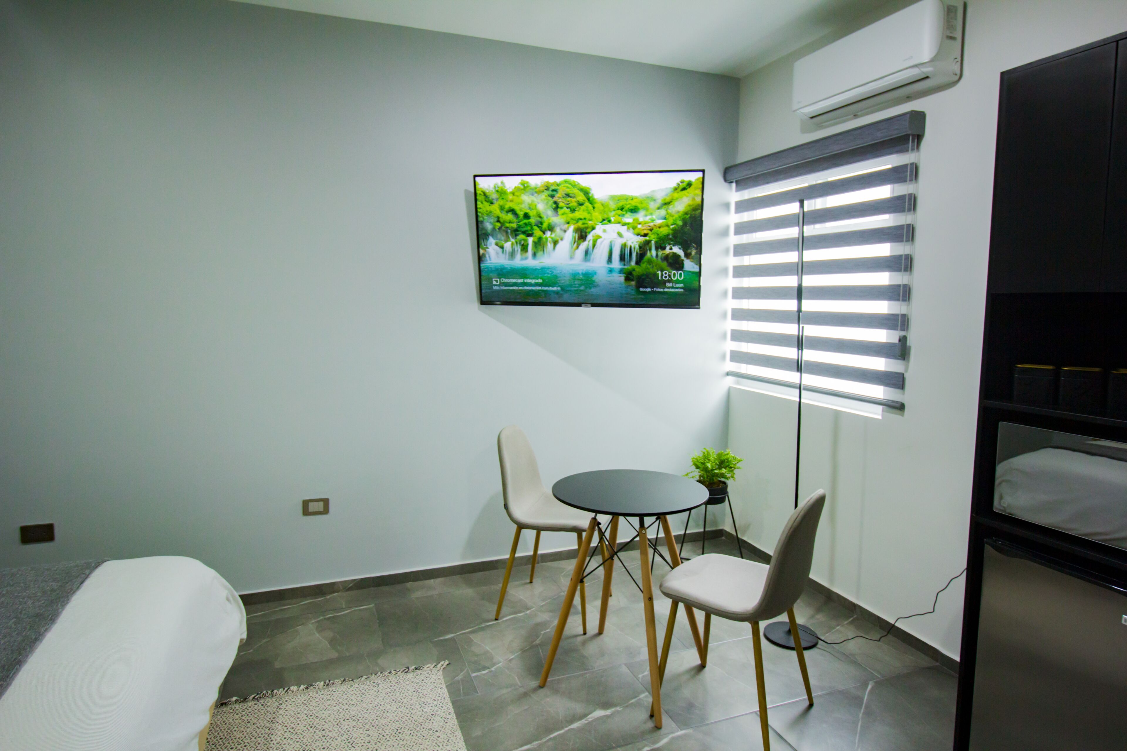 A small room with a bed, a TV mounted on the wall displaying a nature scene, a small table with chairs, and a window with blinds.