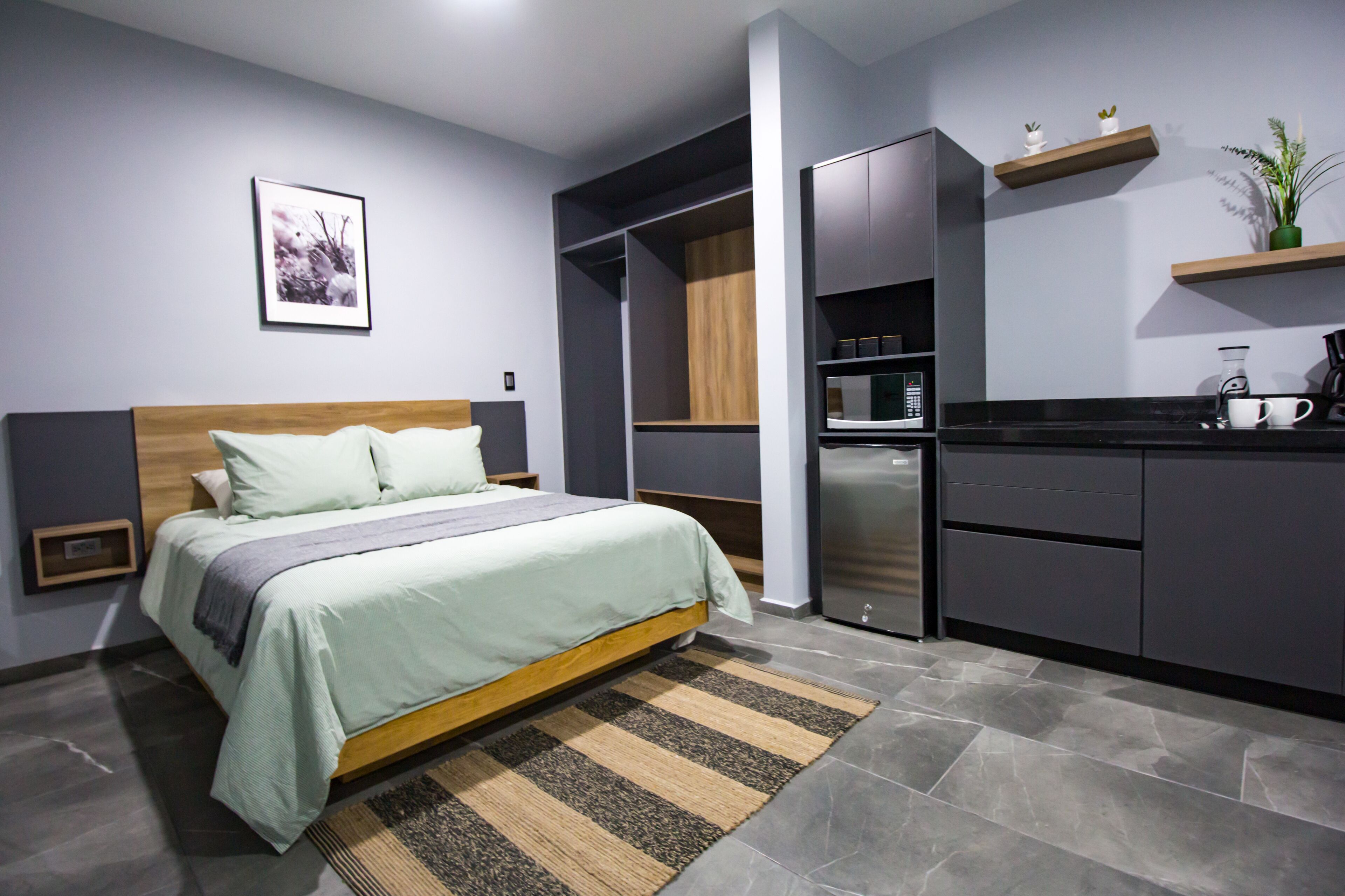 A modern bedroom with a bed, a built-in wardrobe, a kitchen area, and a small shelf with decorative items.