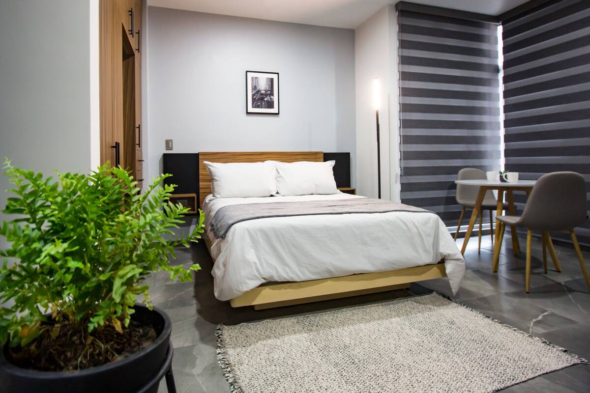 Un dormitorio moderno con cama, mesita de noche, planta en maceta y vista a un comedor.