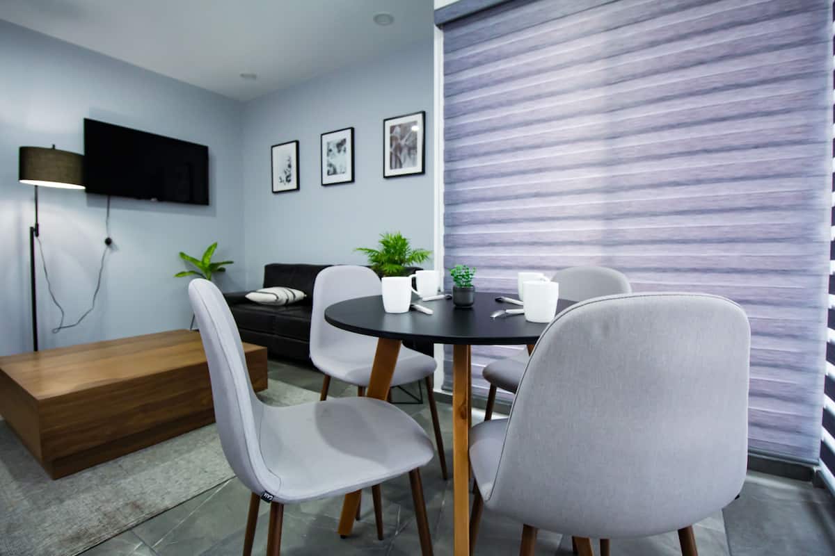 Una sala de estar moderna con una mesa redonda, sillas grises, un sofá, un televisor de pantalla plana montado en la pared y una ventana con persianas.