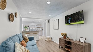 A modern living room with a blue sofa, wooden herringbone-patterned console, and a flat-screen TV displaying a golf scene. - Carriage Manor- Right next to the  Fountain of Youth & Beach.
Pets are welcomed! (St. Augustine)