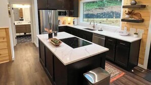 A modern kitchen with a central island, black cabinets, a stainless steel refrigerator, and a large window with a view of greenery. - Modern Mountain Getaway with hot tub & fire pit 10 minutes to Helen (White County)