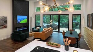 A modern living room with a TV, sofa, dining table, and a large window overlooking a deck. - Modern Mountain Getaway with hot tub & fire pit 10 minutes to Helen (White County)