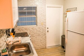 A compact kitchen with a sink, a window with blinds, a refrigerator, and a door.