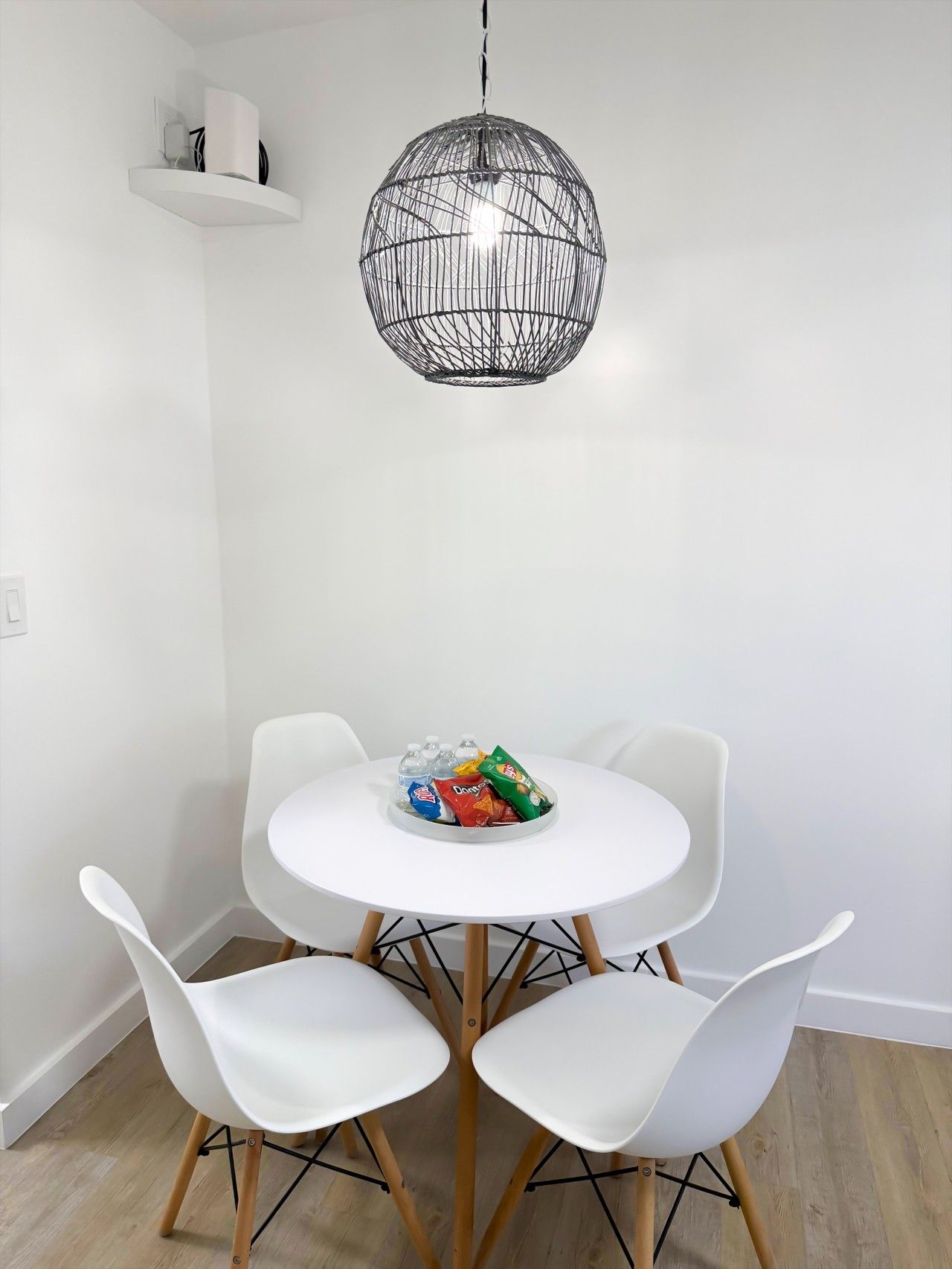 A dining area with a round table, four white chairs, a hanging light fixture, and a small shelf with a white object.