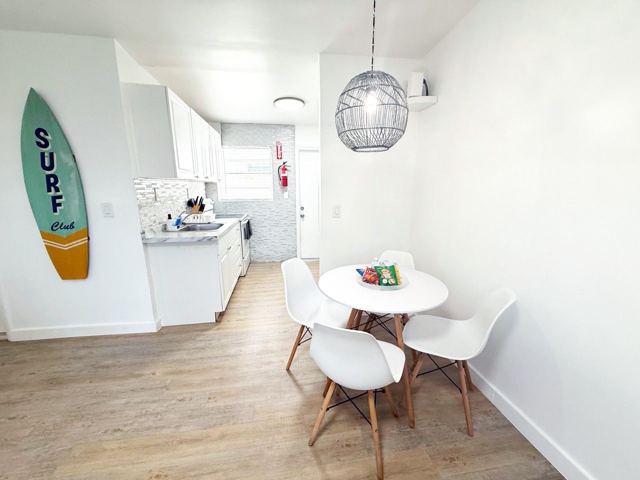 Una cocina moderna con armarios blancos, una zona de comedor con sillas blancas y una decoración de tabla de surf en la pared.