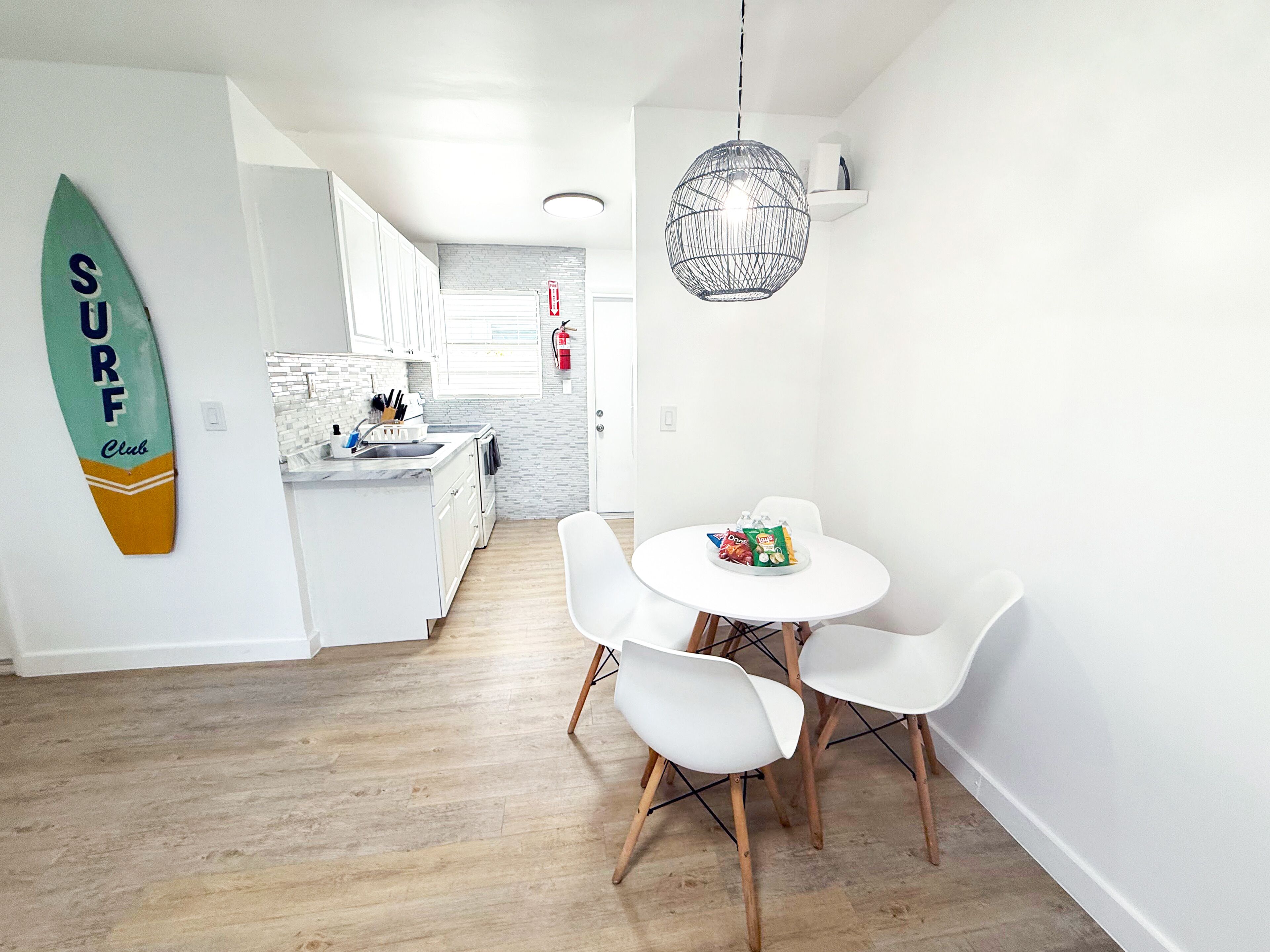 Una cocina moderna con armarios blancos, una mesa redonda con sillas blancas y un adorno de tabla de surf en la pared.