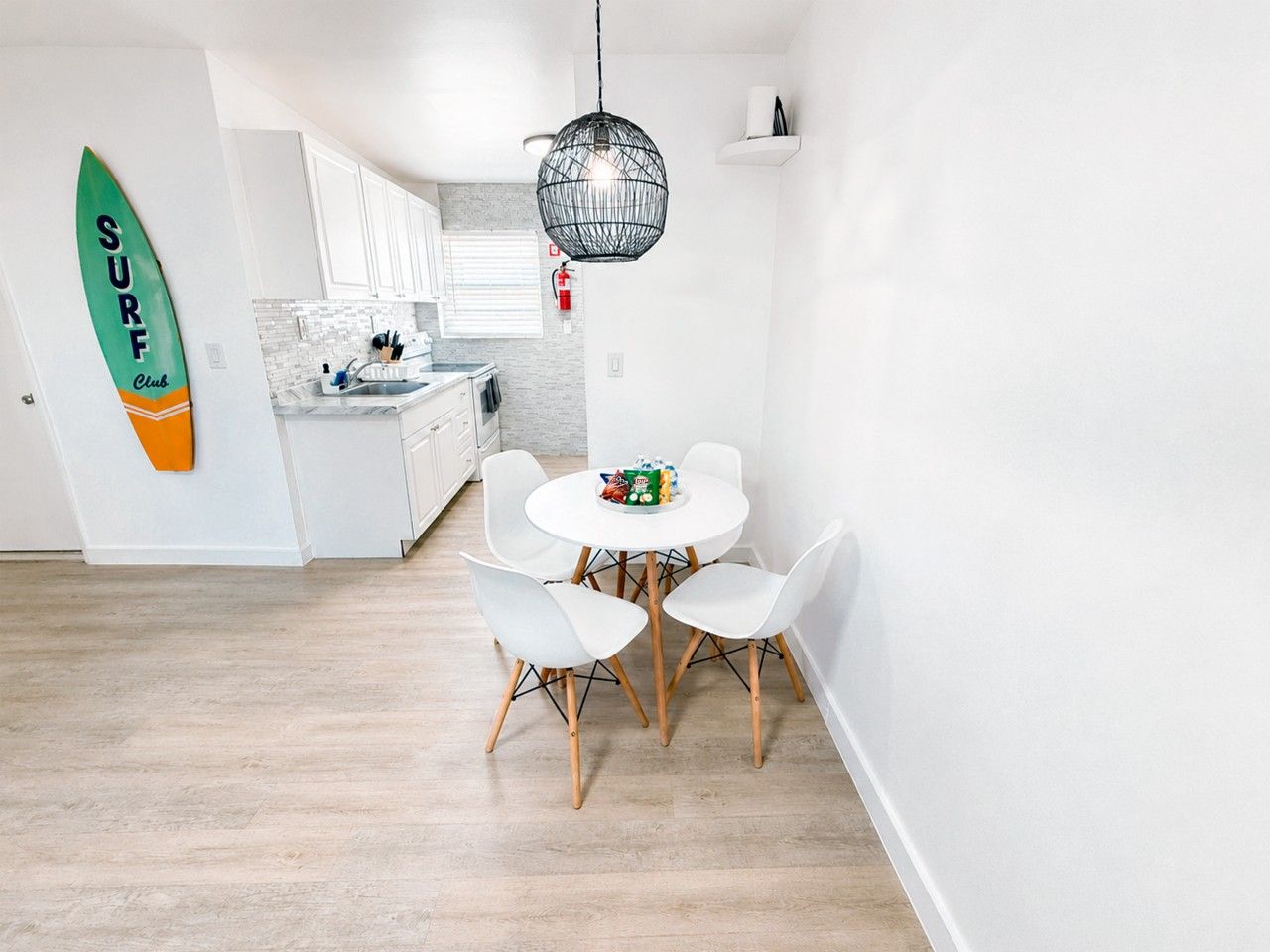 Una cocina moderna con armarios blancos, una mesa de comedor con sillas y un detalle decorativo de tabla de surf.