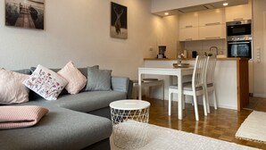 A modern living room with a grey sofa, white dining table, and chairs, a small white side table, and a kitchen area with white cabinets and appliances.