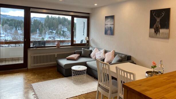 A modern living room with a large window, a grey sofa, a white table, and a wooden dining table.