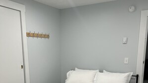 Un lit bien fait, avec du linge de lit blanc et une couverture grise pliée, un porte-manteau en bois et une porte blanche.
