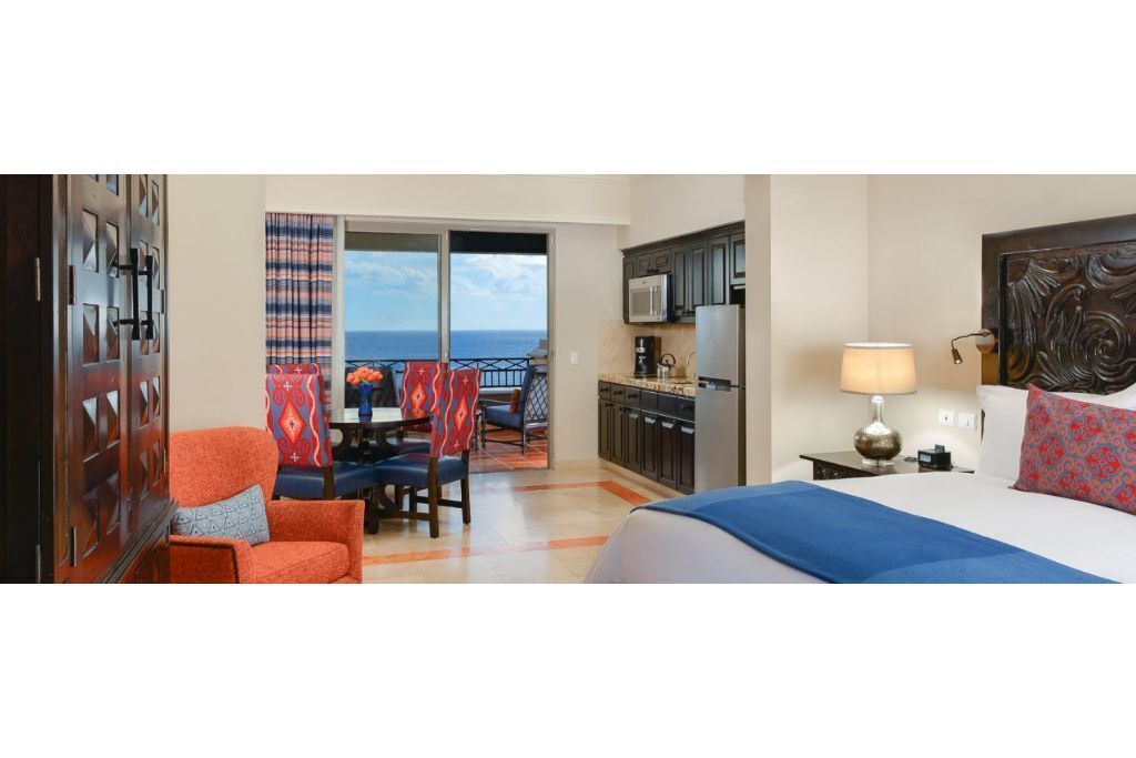 A hotel room with a bed, a desk, a chair, a view of the ocean, and a kitchen area.