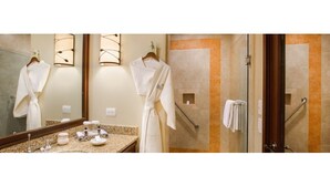 Un baño de hotel con un espejo grande, dos albornoces blancos colgados, una encimera de granito y un box de ducha de vidrio.