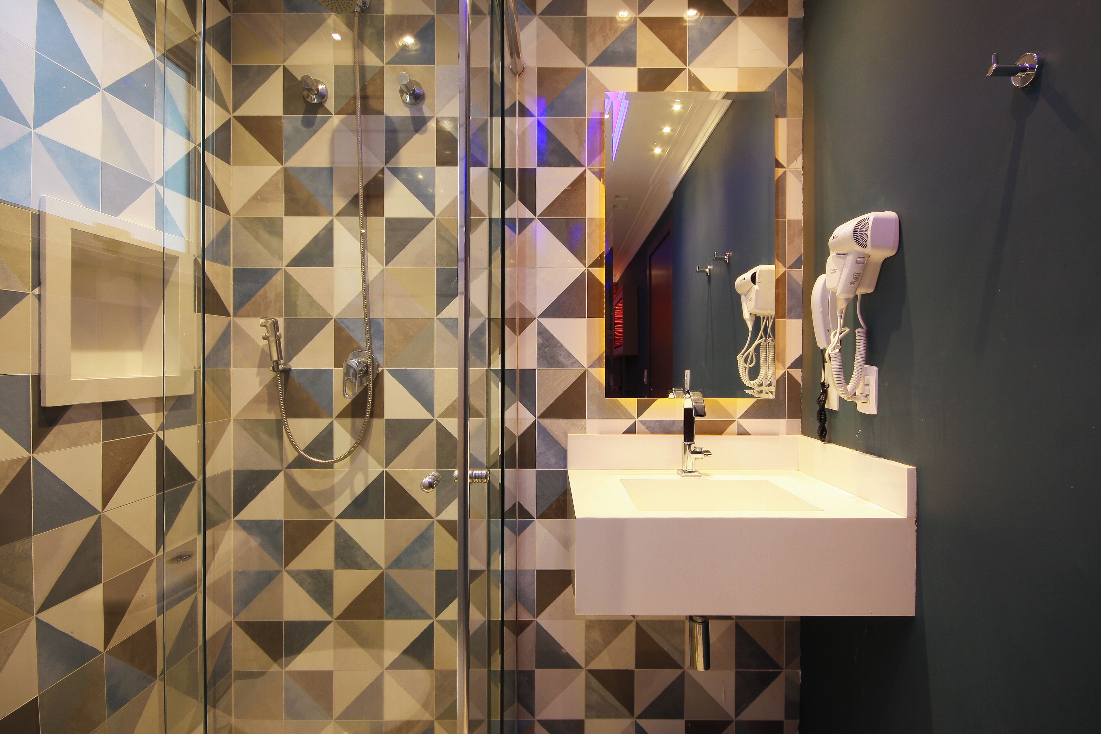 Um banheiro moderno com painel de azulejos geométricos, box de vidro, pia branca e dois secadores de cabelo fixados na parede.
