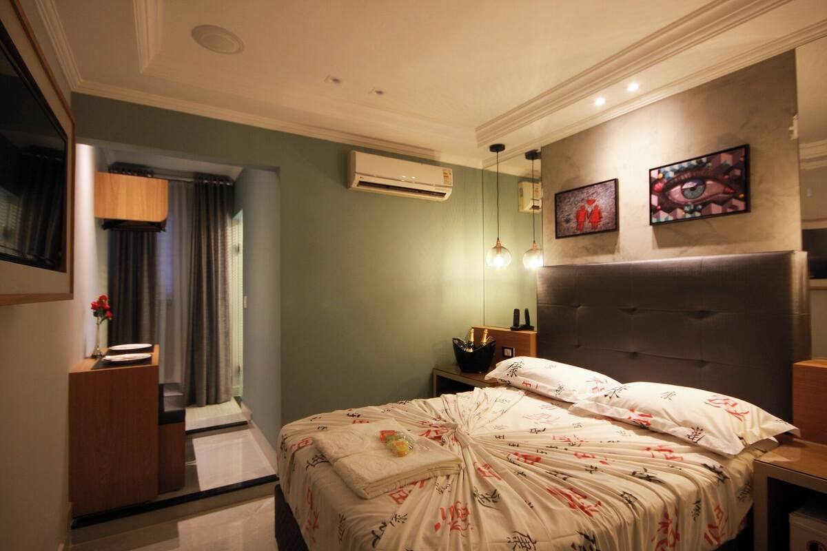 A hotel room with a bed, bedside tables, an air conditioning unit, a mirror, and artwork on the wall.