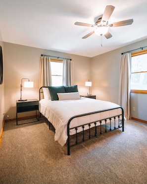 Une chambre à coucher avec un lit, deux tables de chevet, un ventilateur de plafond et une fenêtre avec des rideaux.