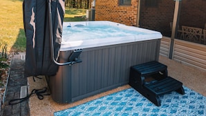 Un bain à remous dans un jardin, avec un bâtiment en briques, une terrasse en bois et un tapis bleu à motifs.