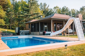 Une piscine avec un toboggan, un patio couvert et une maison en briques située dans un endroit boisé.