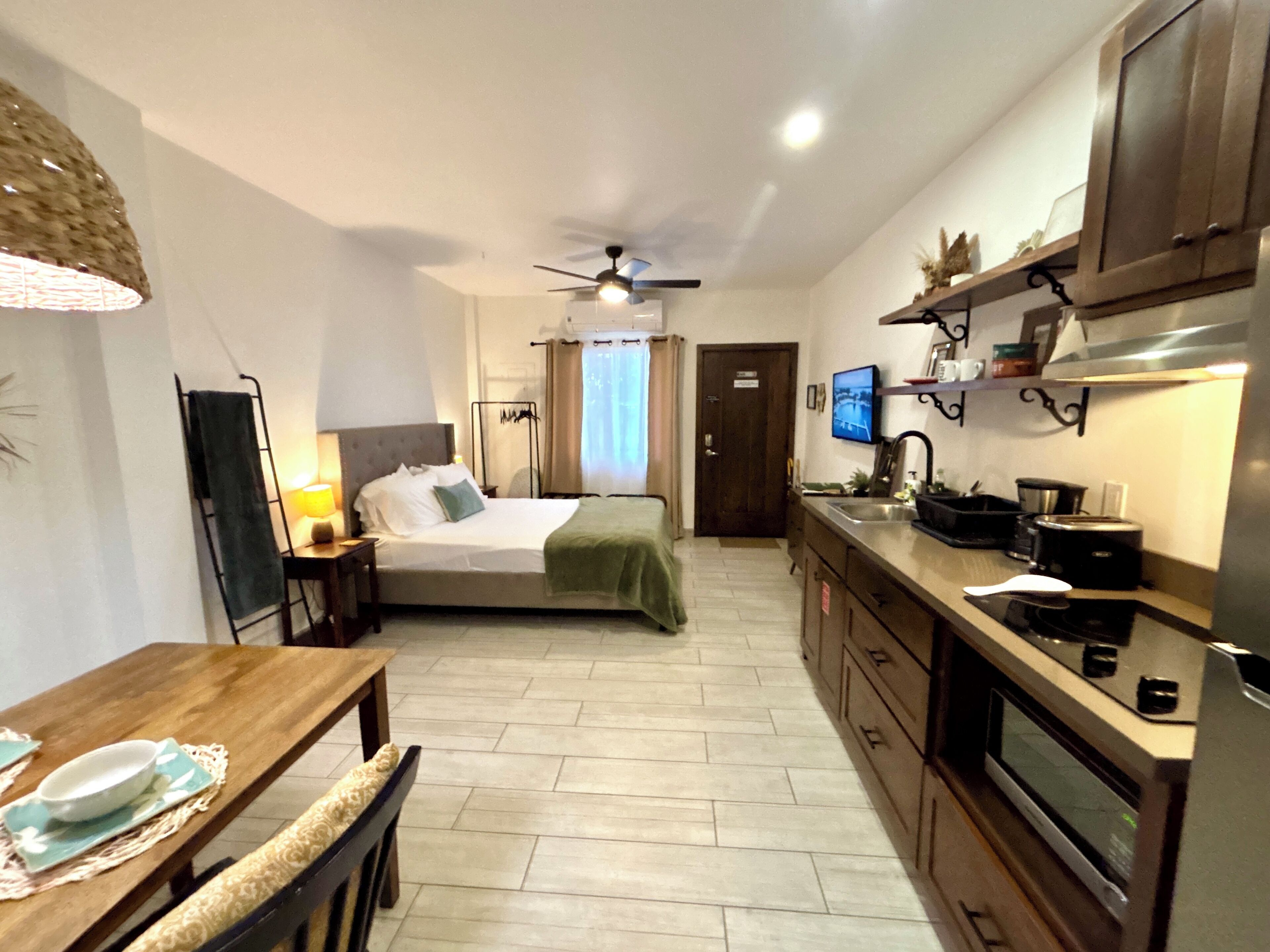 A compact hotel room with a bed, a dining table, a kitchenette, and a ceiling fan.