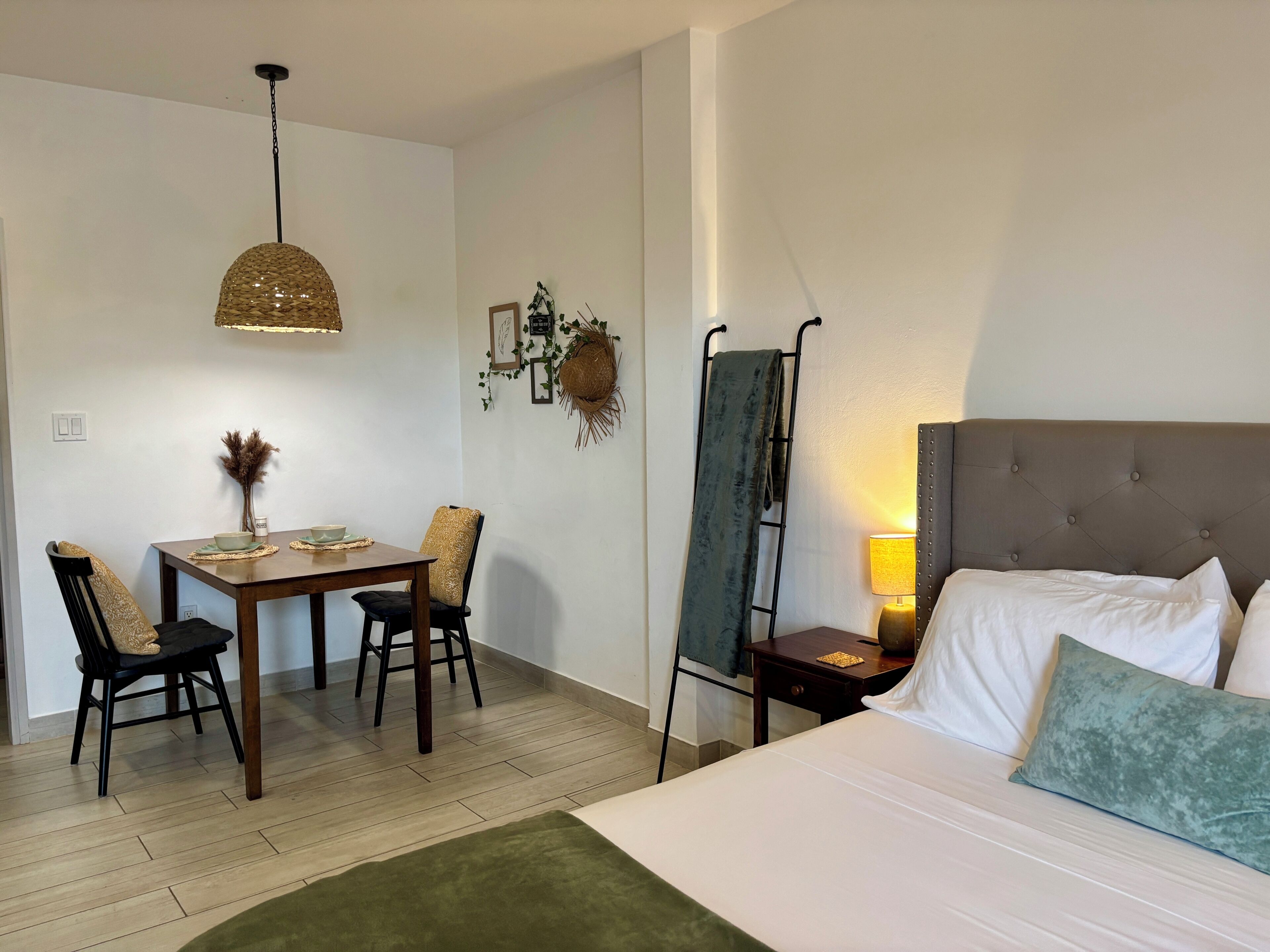 Une chambre d’hôtel comprenant un lit, un bureau avec des chaises, un luminaire suspendu et une étagère fixée au mur.