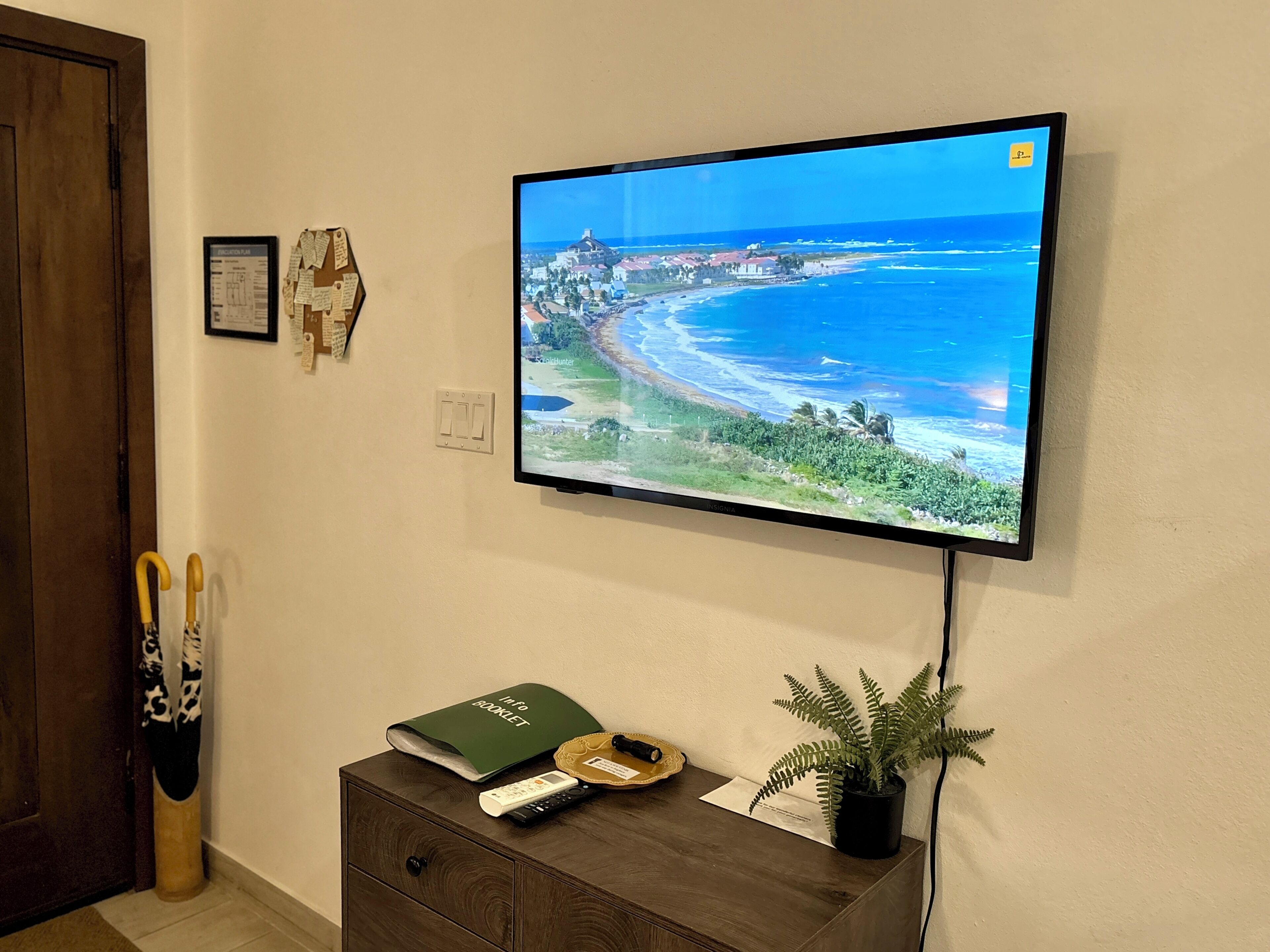 A television mounted on a wall displaying a coastal scene, a wooden cabinet with a remote and a book, and a door with a cane leaning against it.